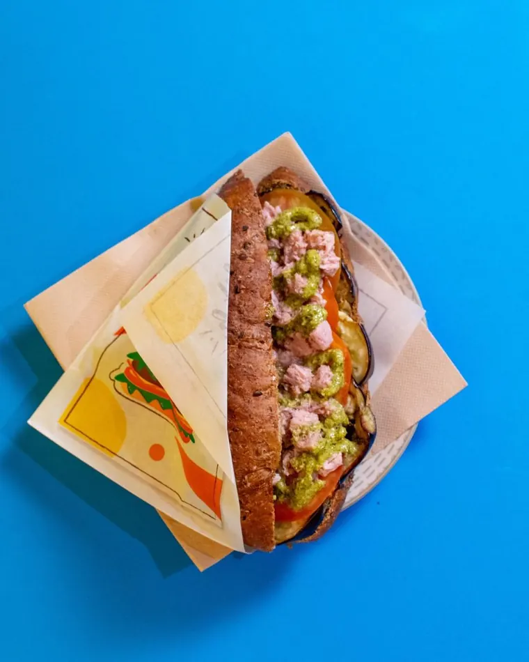 Panino con tonno, melanzane, pomodoro e pesto, visto dall'alto su fondo azzurro
