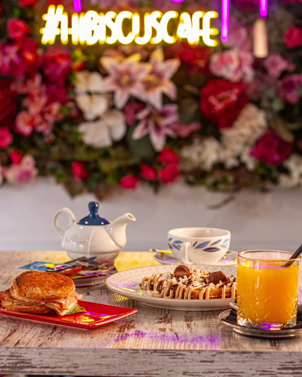Parete di fiori rossi e rosa con l'insegna al neon del locale, davanti a un tavolo con colazione