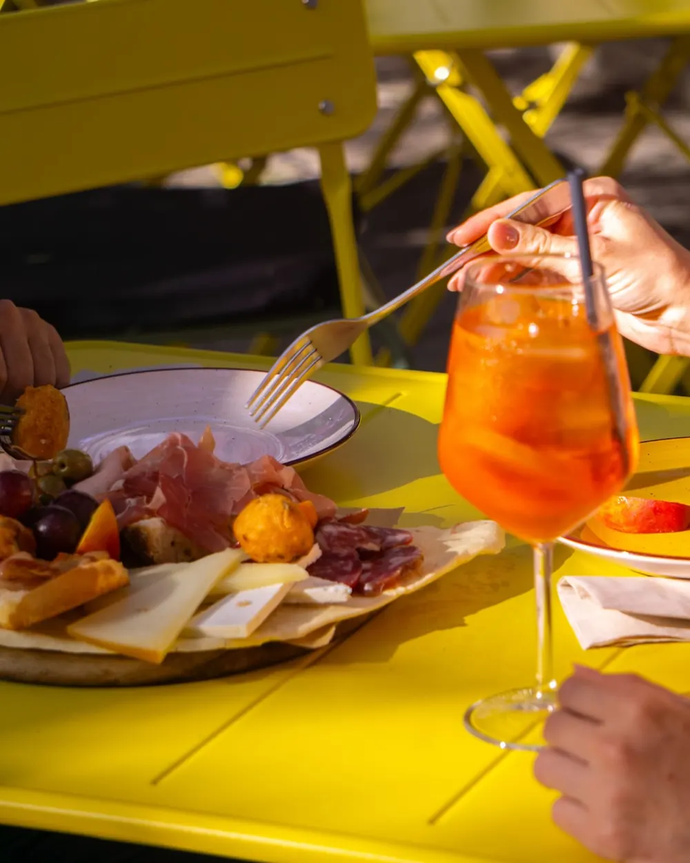 Tagliere di salumi e formaggi con pane carasau e uno spritz, su un tavolino giallo all'aperto